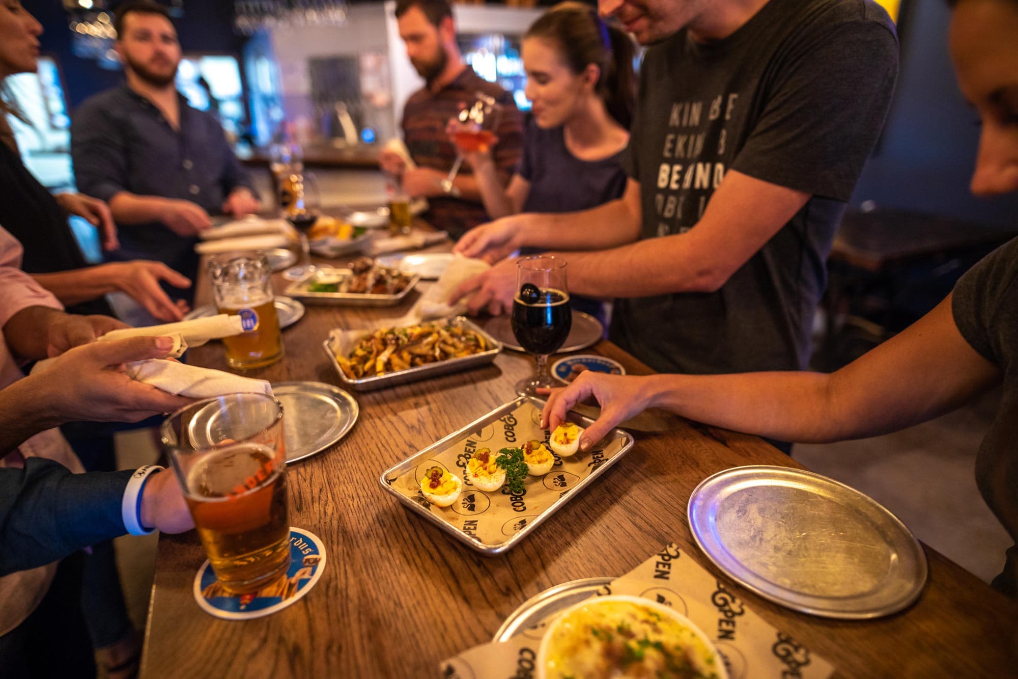 adults gathered around table at Cob & Pen in Lakeland, FL drinking beer and sharing small bite plates of deviled eggs, dip, french fries and wings